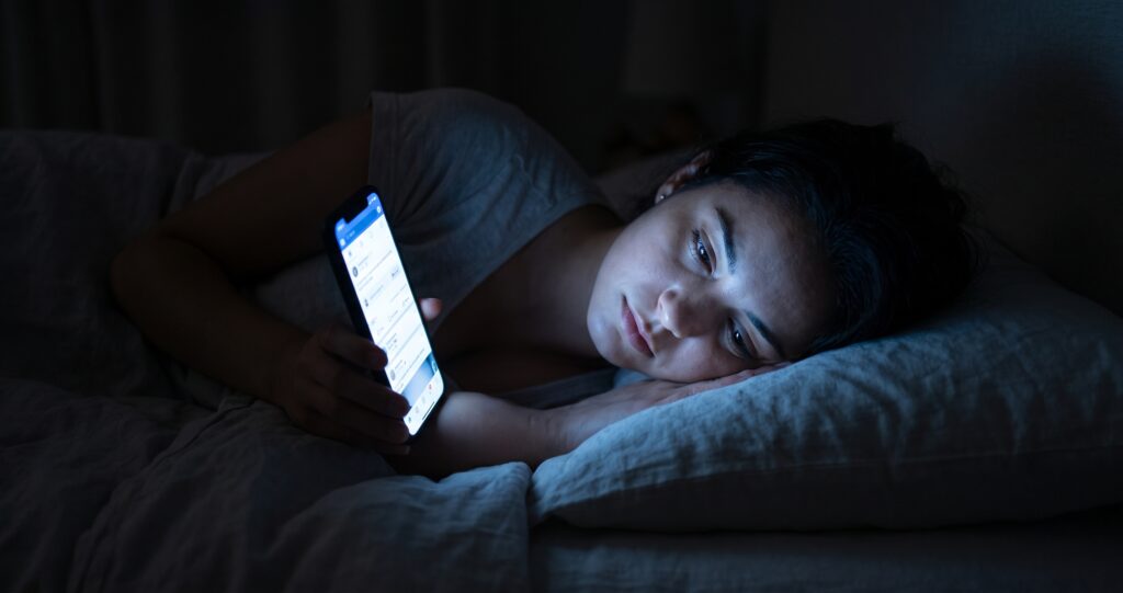 Mulher deitada na cama à noite, no escuro, iluminada apenas pela luz azul da tela do seu smartphone.
